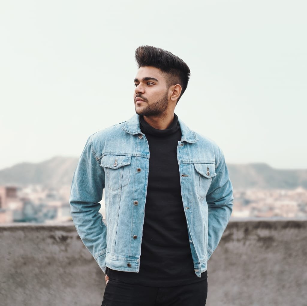 A man wearing a light blue denim bomber jacket over a black turtleneck, standing outdoors with a cityscape and hills in the background.