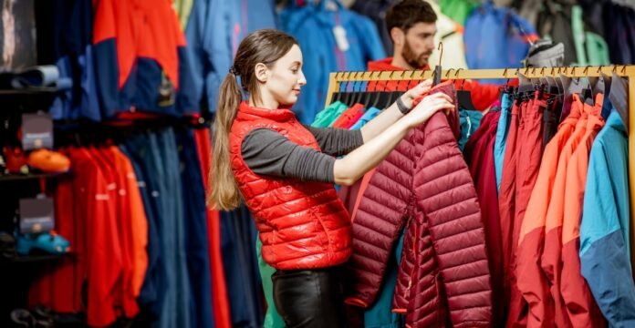 A woman shopping for outdoor jackets in a store, browsing through a rack of colorful insulated jackets while a man nearby looks at other clothing.