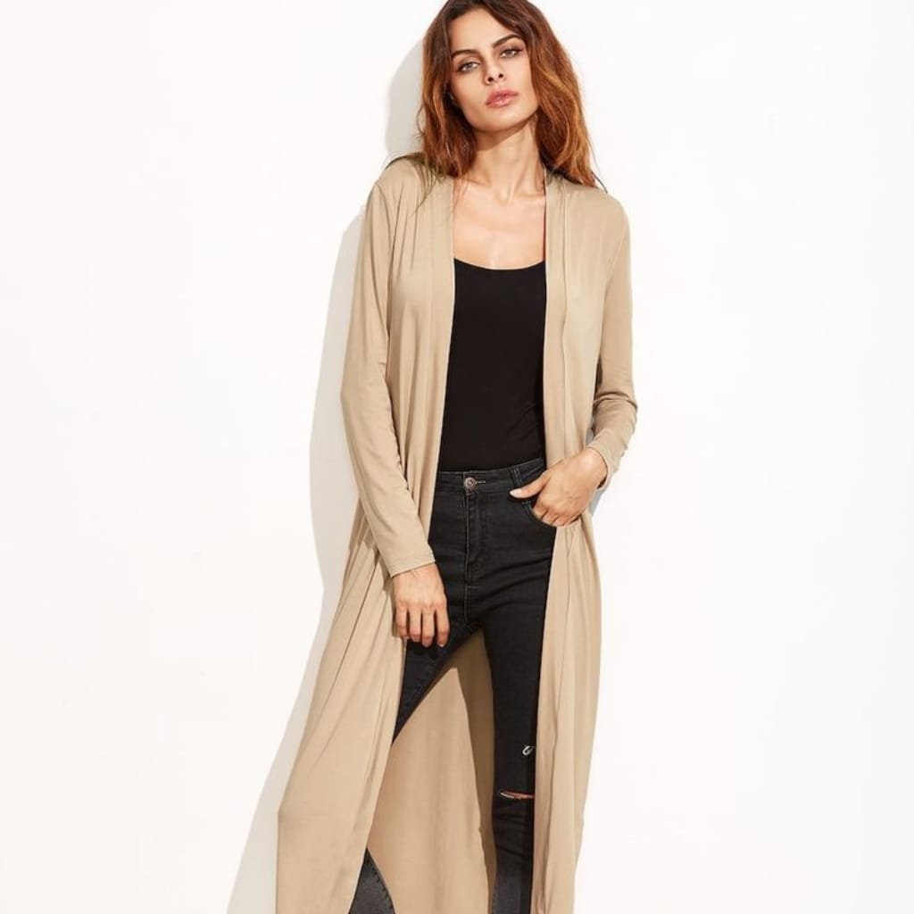 A woman with wavy auburn hair wearing a long, beige duster coat over a black top and black ripped jeans, standing against a plain white background.
