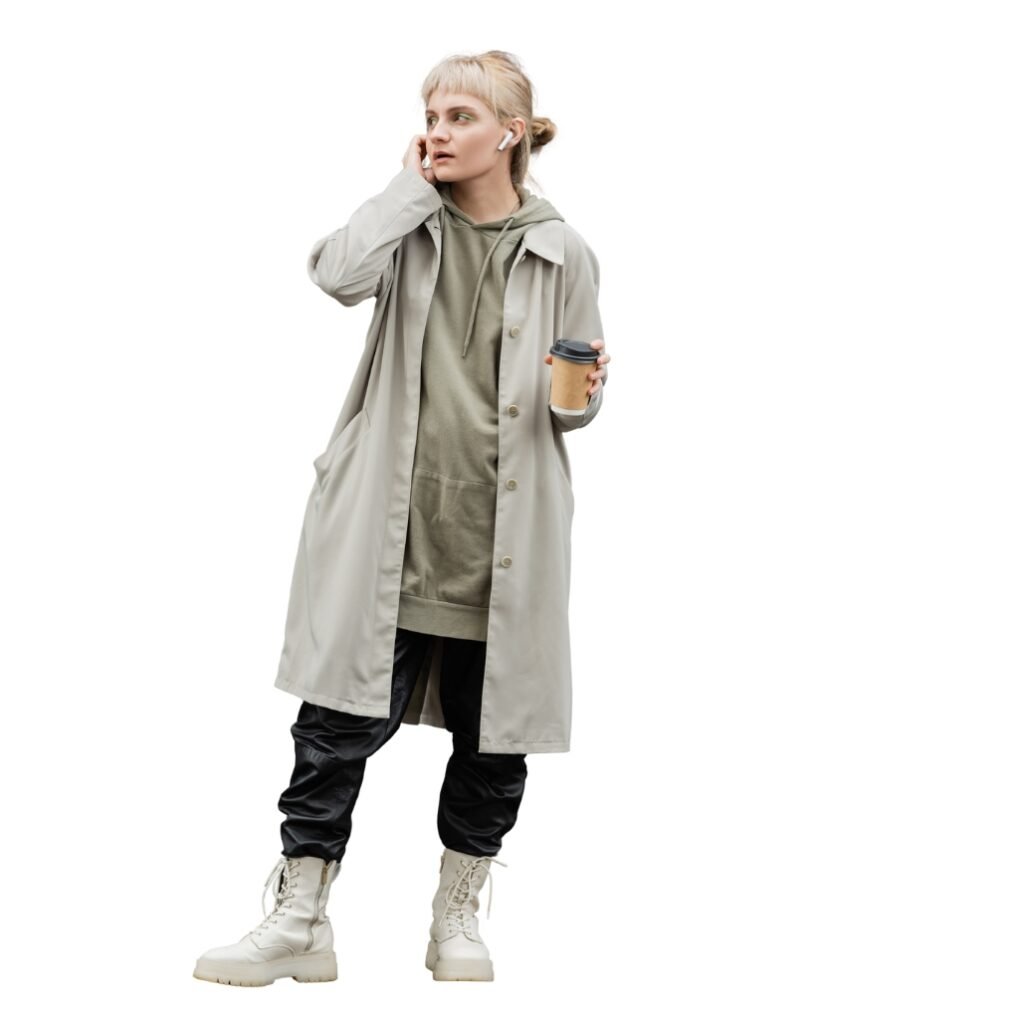 A young woman dressed in casual, layered clothing, holding a coffee cup, and wearing a long windbreaker jacket, earbuds, and hiking boots, standing against a white background.