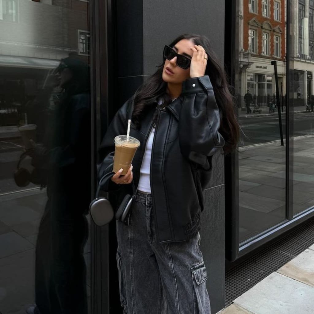 Stylish Black Jackets: The Wardrobe Essential for Every Season 1 A woman standing outside a building, wearing a stylish black leather jacket, oversized sunglasses, and casual black cargo jeans, holding an iced coffee with a straw. She has long, dark hair and a confident, relaxed pose.