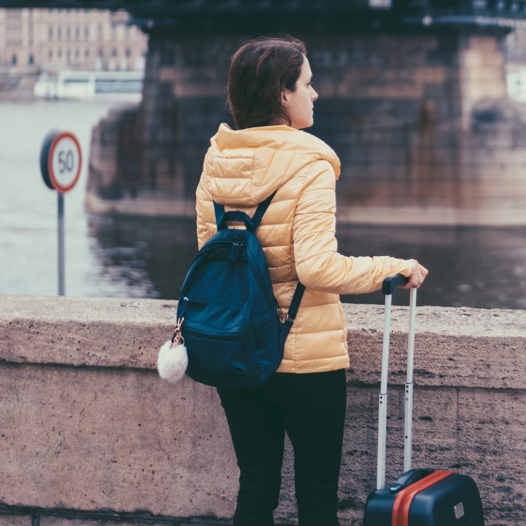 A woman standing by a river, wearing a beige quilted jacket, with a backpack on her shoulders and pulling a rolling suitcase.

