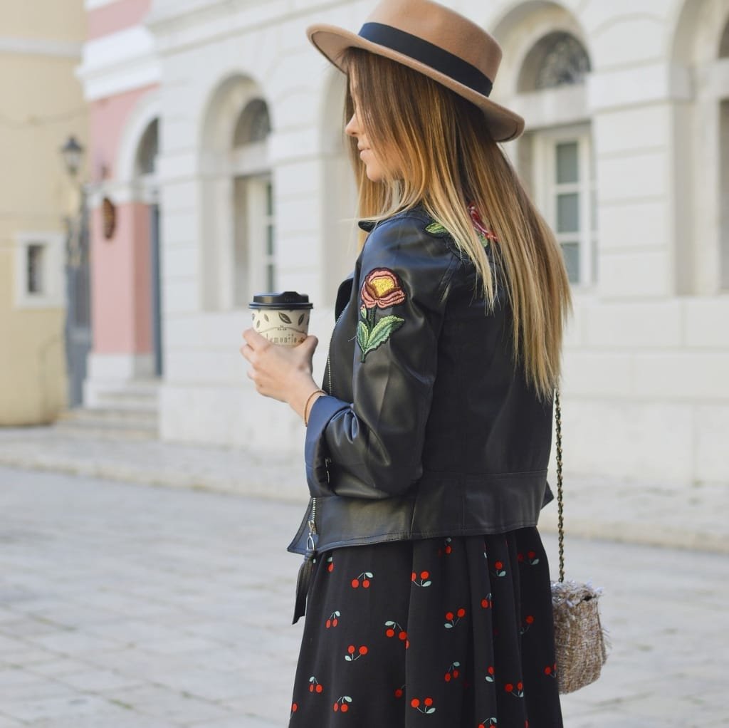 A woman standing outdoors, holding a coffee cup, wearing a black embroidered bomber jacket with floral designs on the sleeve.