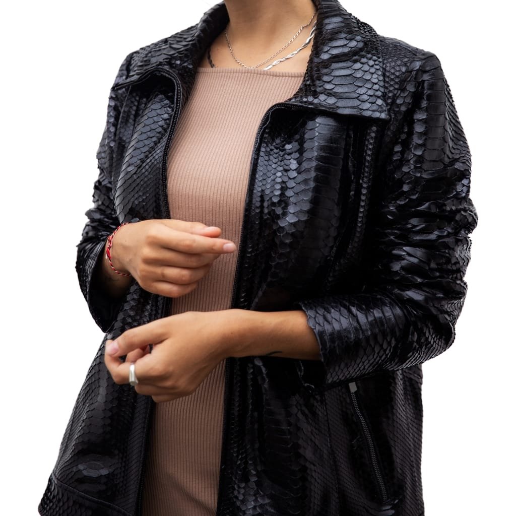 A woman wearing a shiny black snakeskin-patterned jacket over a beige ribbed dress, holding her hands in front of her, with layered necklaces and a ring on her finger, suitable for an evening out.

