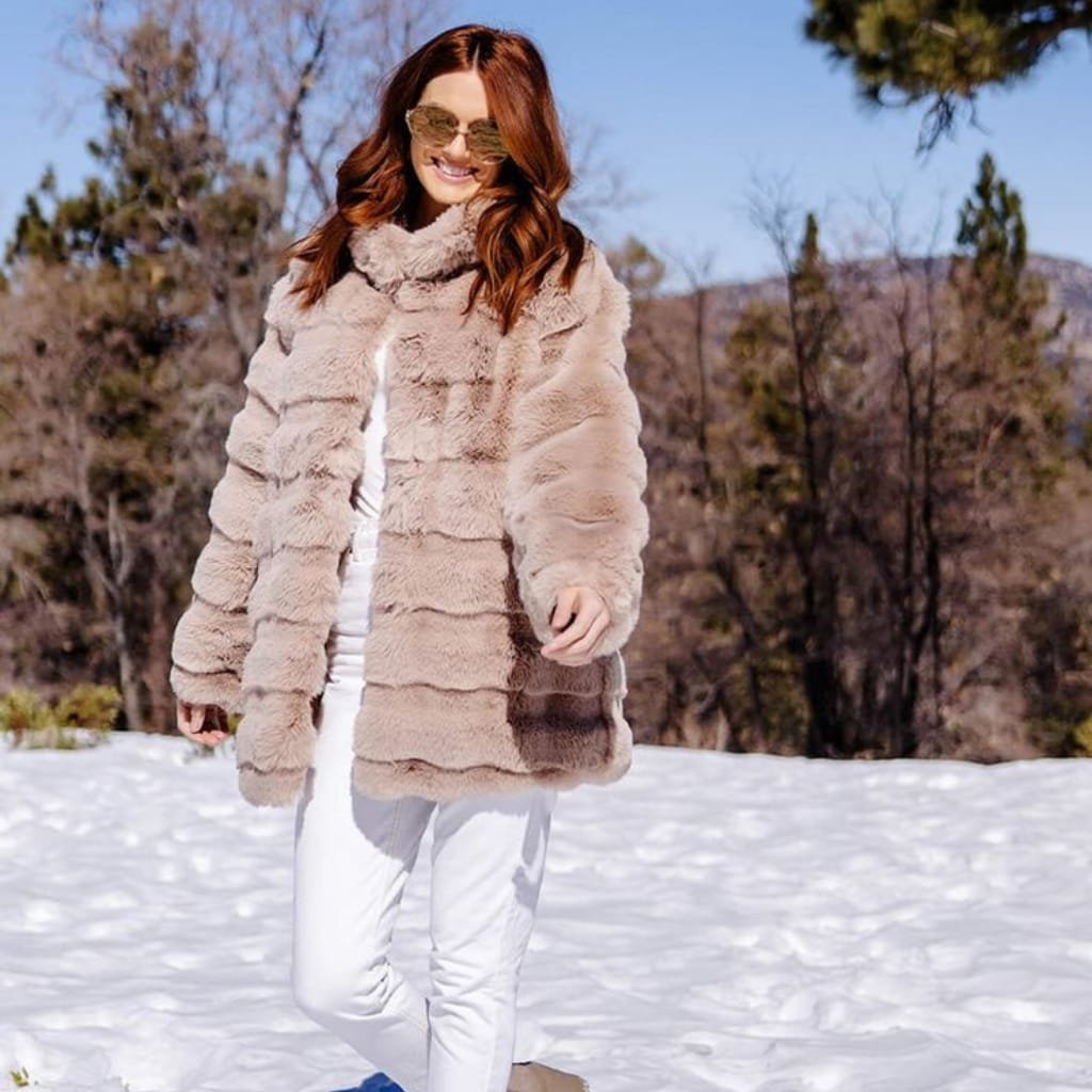 A woman walking outdoors in a snowy landscape, wearing a plush, beige faux fur coat, white pants, sunglasses, and a bright smile, enjoying a sunny winter day.
