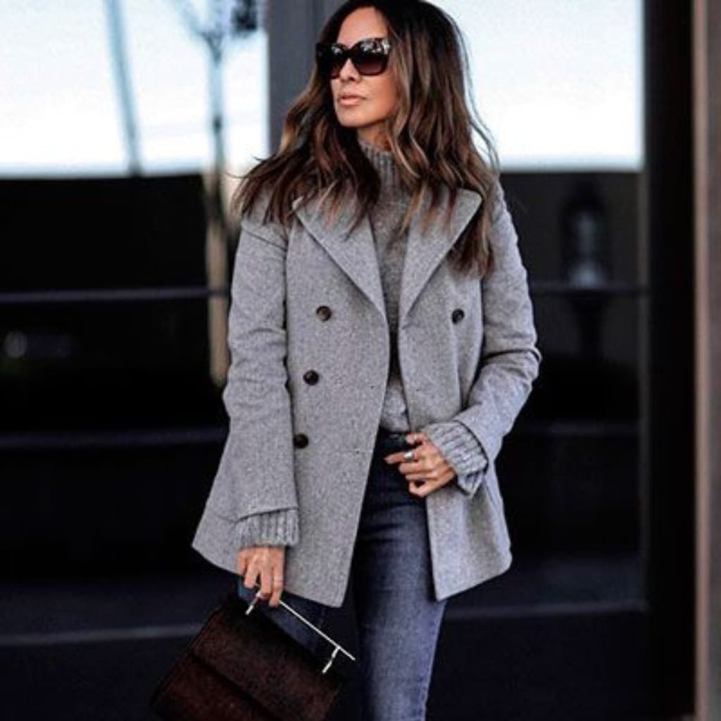A stylish woman wearing a light gray pea coat, dark jeans, and sunglasses, carrying a dark handbag, standing outdoors on a fall day