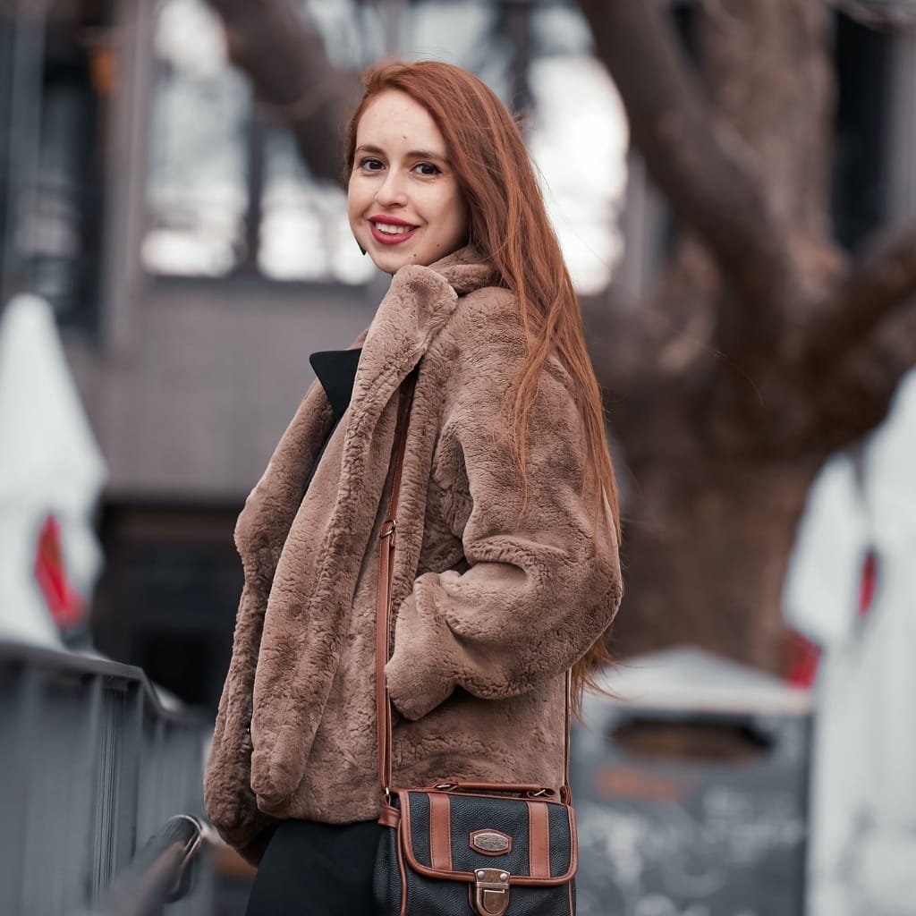 A young woman with long red hair smiling outdoors, wearing a plush, brown faux fur coat and carrying a black and brown handbag.

