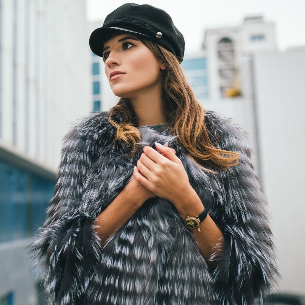 A woman wearing a luxurious faux fur coat, standing outdoors in an urban setting, with her hands gently clasped near her chest and a contemplative expression.