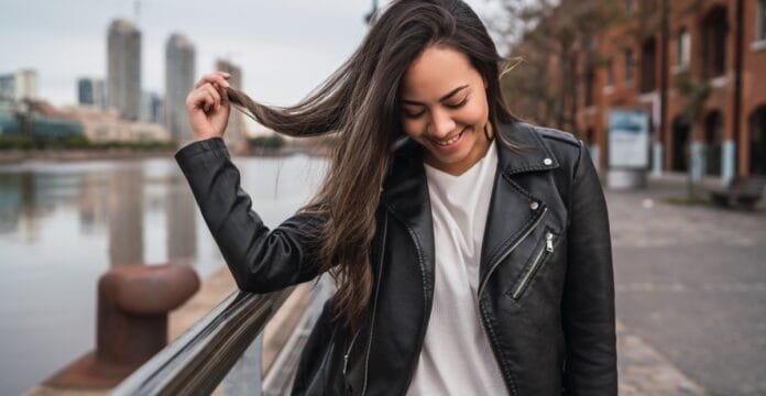A young woman with long, dark hair wearing a black leather jacket, smiling and playfully holding her hair while standing outdoors near a river with a cityscape in the background.