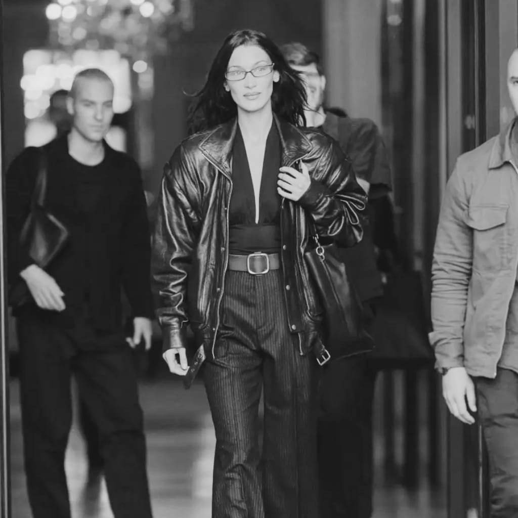 A black and white photo of a woman with dark hair and glasses walking through a city street, dressed in a leather jacket and high-waisted pinstripe trousers. She appears confident and composed, with other pedestrians around her.