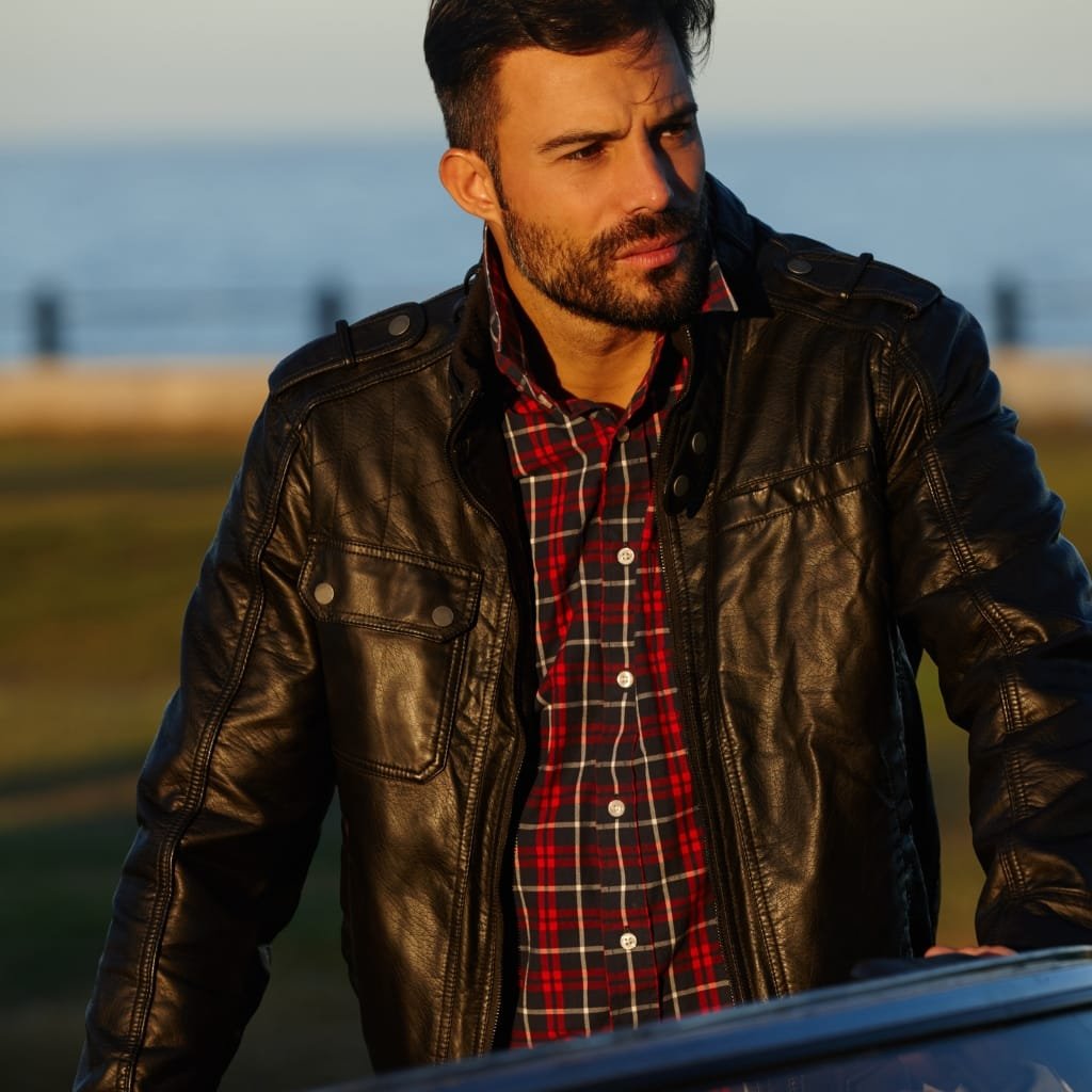 A man with dark hair and a beard wearing a black leather shirt jacket over a red and black plaid shirt, standing outdoors near a waterfront at sunset.

