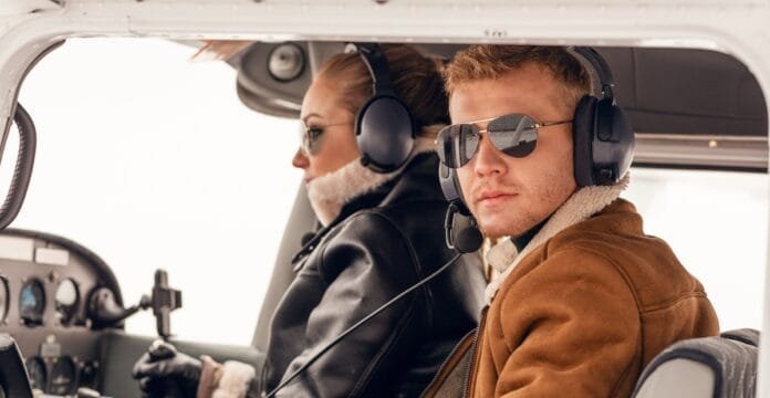 A man and woman sitting in the cockpit of an aircraft, wearing flight jackets and aviation headsets, with the woman at the pilot's controls and the man looking towards the camera.