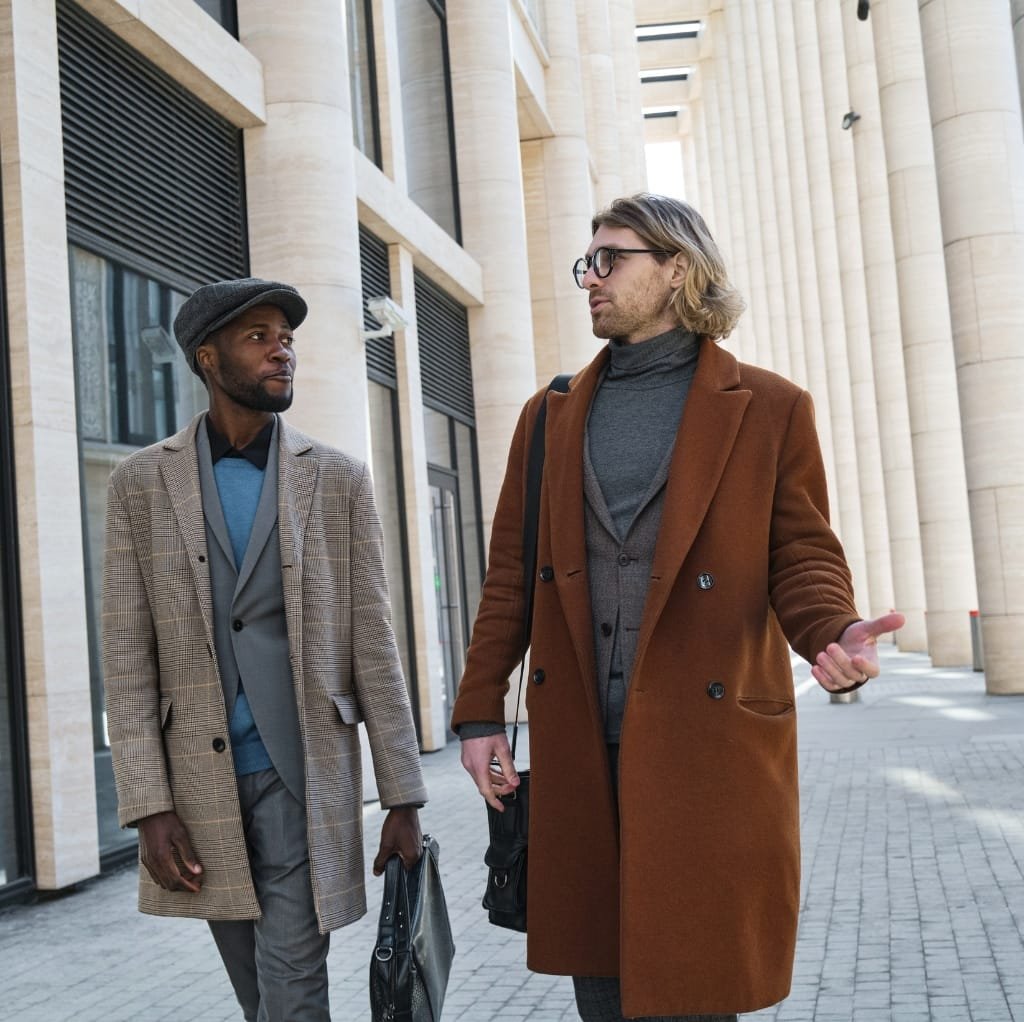 Two men walking and talking outside a modern building, dressed in stylish wool work coats layered with sweaters and shirts, demonstrating smart layering fashion.