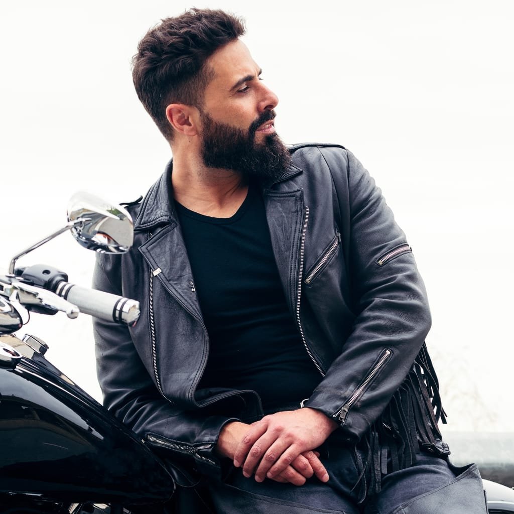 A man with a full beard and styled hair wearing a black leather jacket over a black t-shirt, sitting on a black motorcycle against a plain white background.