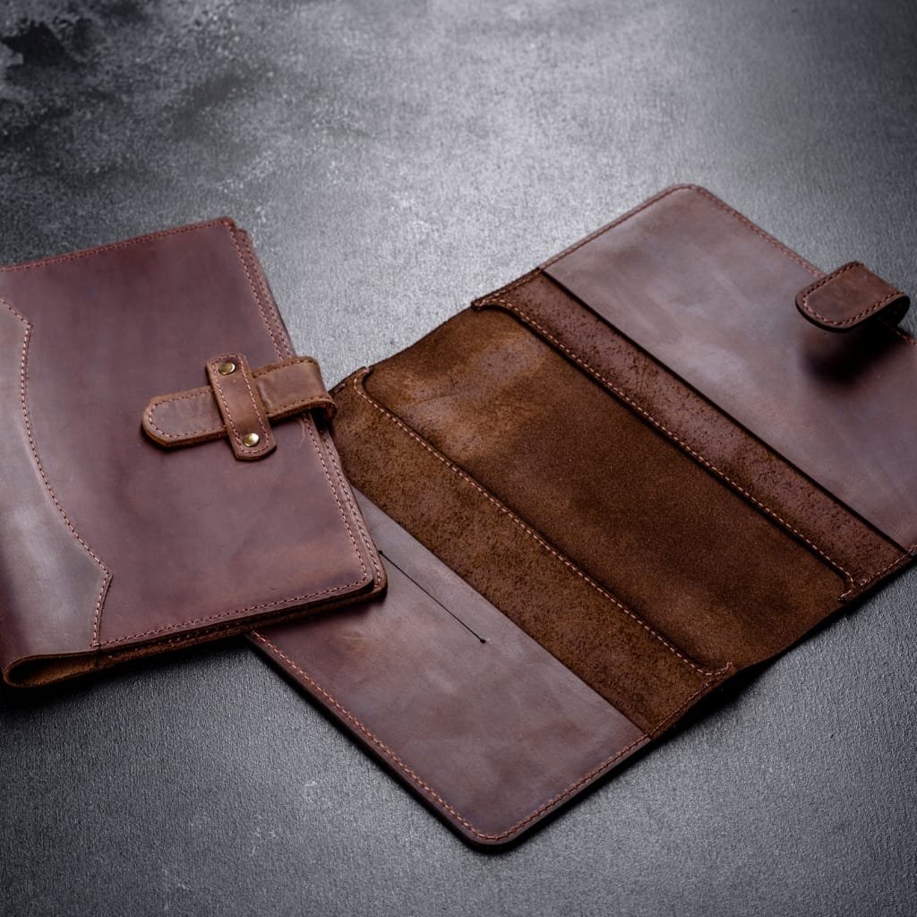 A brown leather portfolio folder with a snap strap, lying open on a dark textured surface.