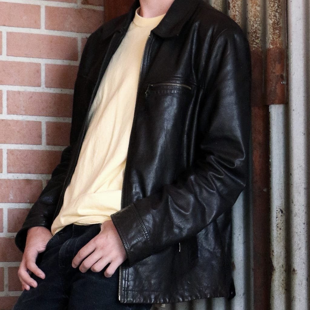 A person wearing a black full or top-grain cowhide leather shirt jacket, standing against a brick wall and a rusted metal pipe.