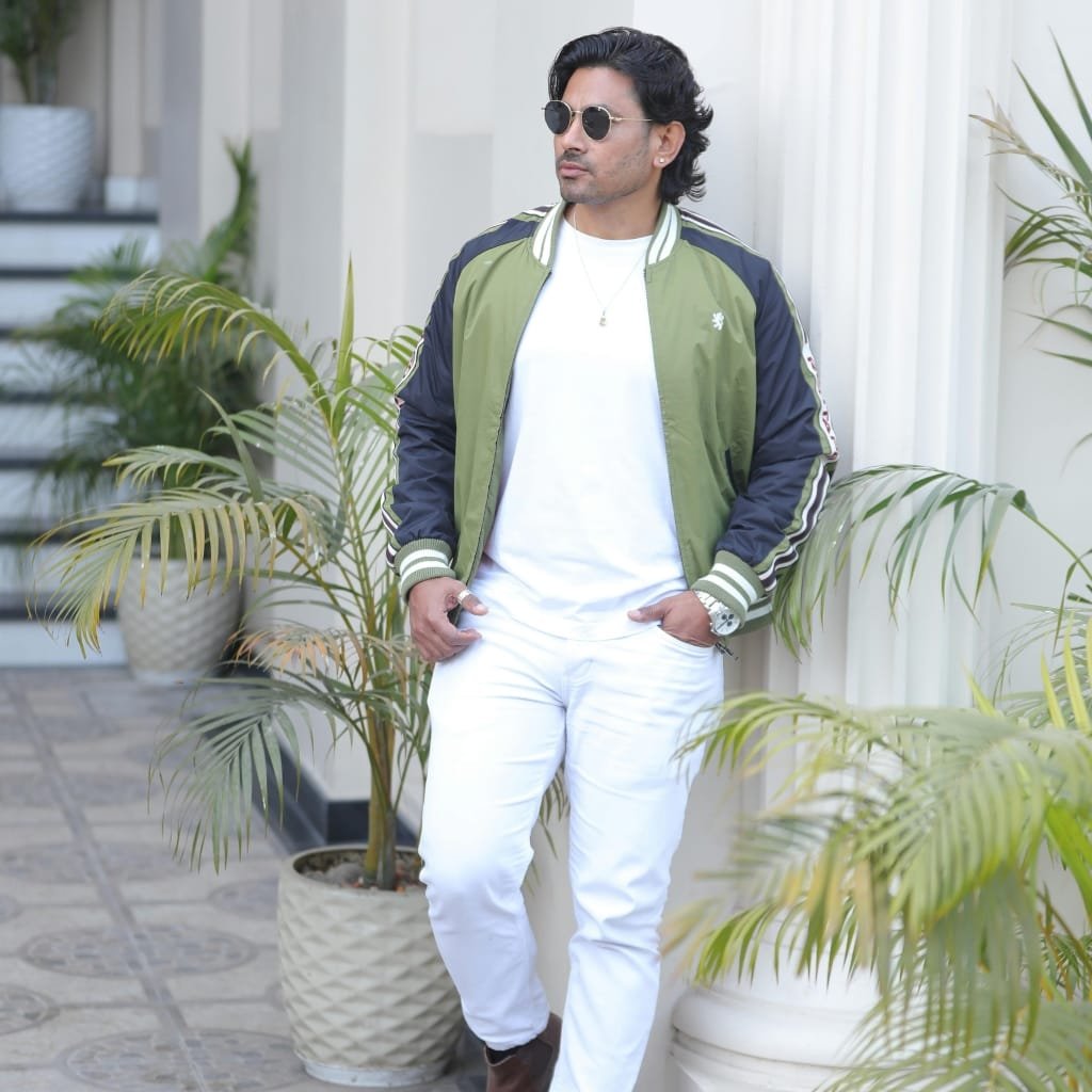 A man wearing a stylish green and navy bomber jacket over a white t-shirt, paired with white jeans, standing beside potted plants against a light backdrop. He has black wavy hair, sunglasses, and accessorizes with a watch and jewelry.

