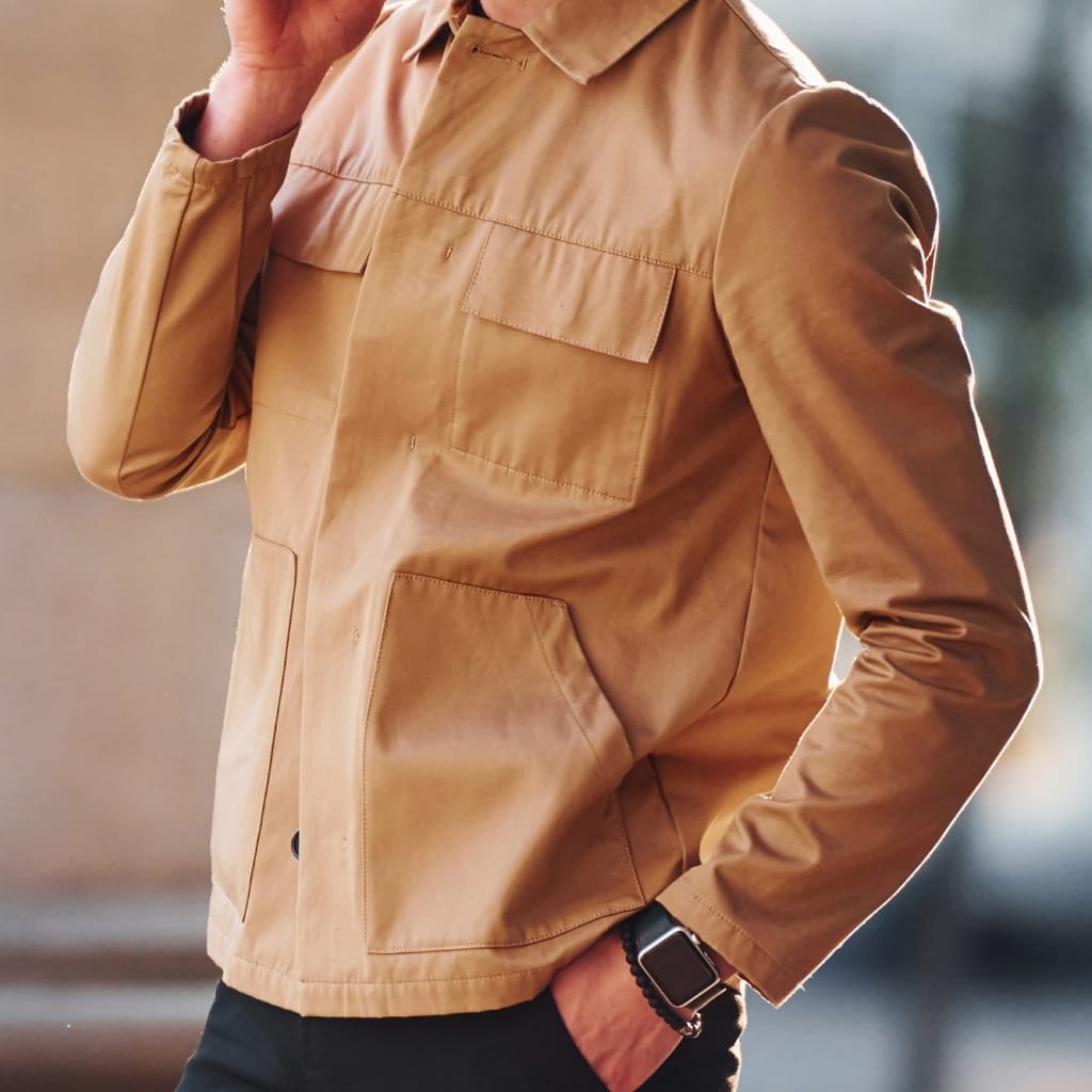 A person wearing a tan goatskin leather shirt jacket with large front pockets, rolled-up sleeves, and a collar.

