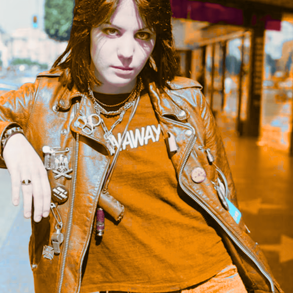 A young person with dark, shoulder-length hair, wearing a brown leather jacket and multiple necklaces, posing on a city street with a confident expression.