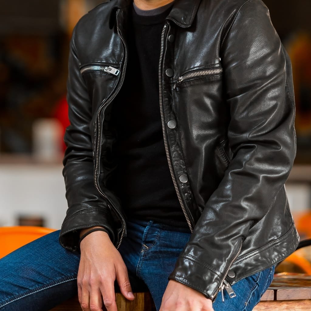 Close-up of a person wearing a black leather jacket, sitting with one hand resting on their knee. The jacket features metal zippers and snap buttons, set against a blurred indoor background.