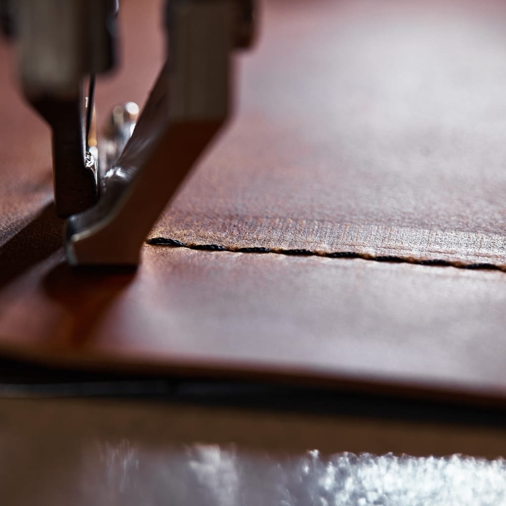 Close-up image of a sewing machine stitching through a piece of full grain leather, highlighting the texture and natural imperfections of the material.