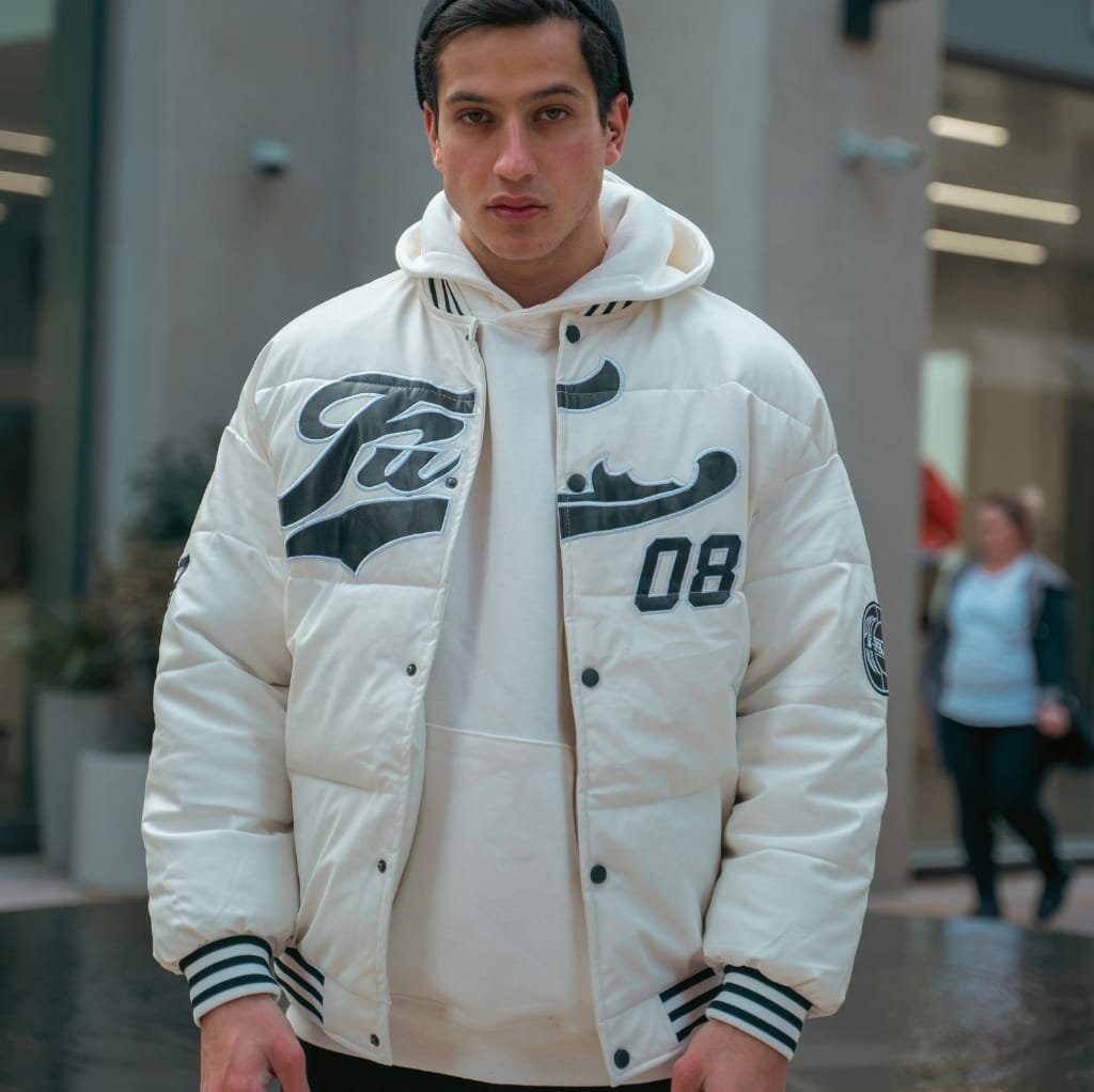 A young man wearing a white quilted bomber jacket with bold black graphics, standing in an urban shopping area.