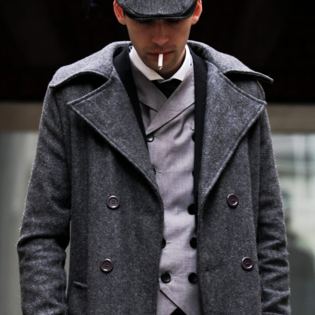 A man wearing a stylish dark gray pea coat over a gray vest and tie, with a cigarette in his mouth, standing outdoors.