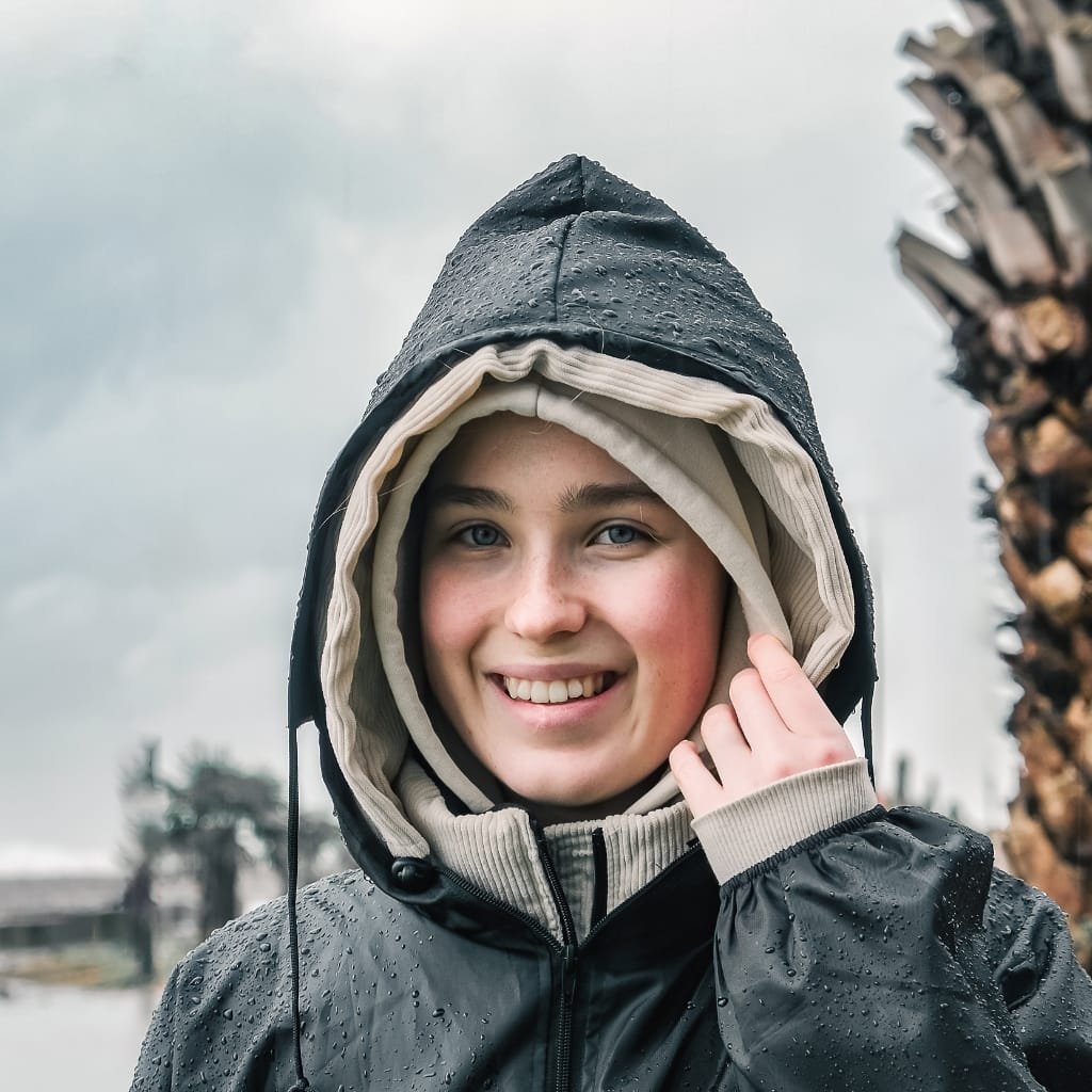 A young woman smiling outdoors in rainy weather, wearing a rain jacket with the hood up over layered clothing, with raindrops visible on the jacket.