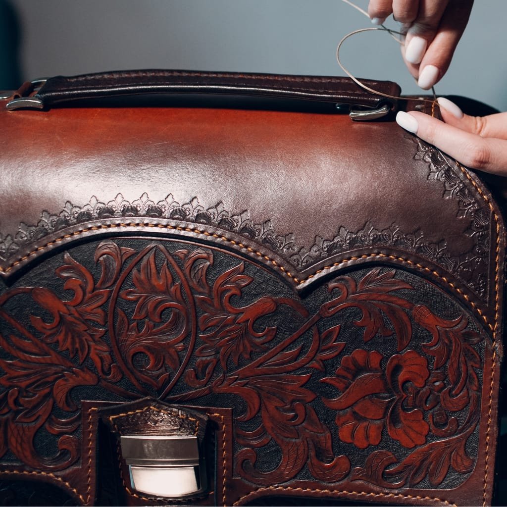 A close-up of a person sewing or repairing a curly leather trim on a polished, dark brown full grain leather bag with intricate floral embossing.