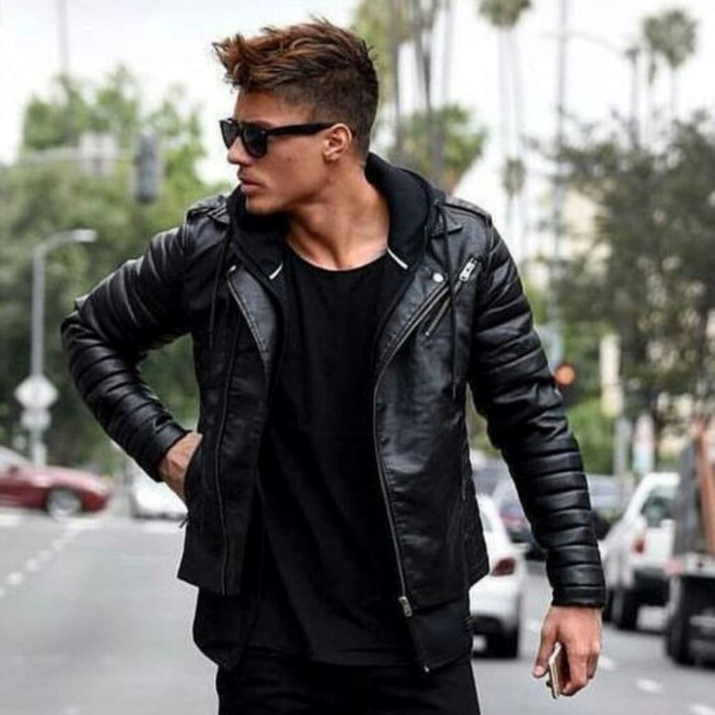 A stylish young man wearing a fitted black leather jacket, sunglasses, and black clothing, standing outdoors on a street with trees and cars in the background.

