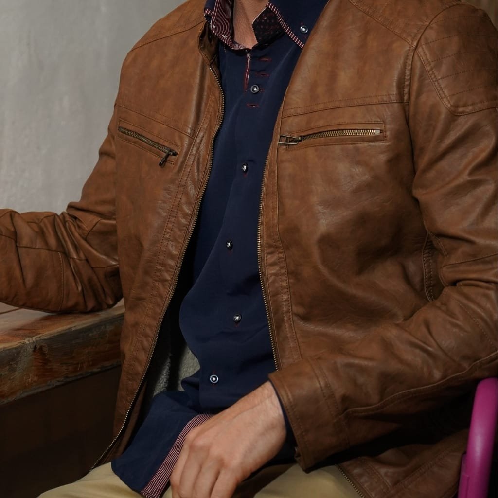 A man wearing a brown lambskin leather jacket over a navy blue shirt, seated with one arm resting on a wooden surface.