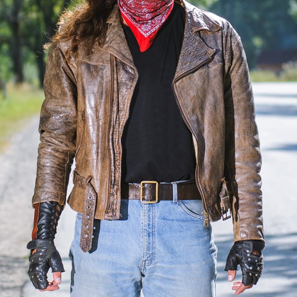 A person dressed in a brown leather motorcycle jacket, black gloves, black t-shirt, and light blue jeans, standing outdoors on a sunny day.

