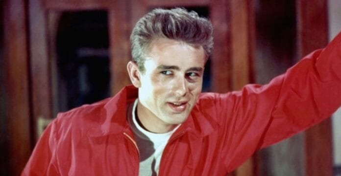 A young man with light brown hair, wearing a red jacket over a white shirt, is raising one arm and making a playful, slightly mischievous expression.