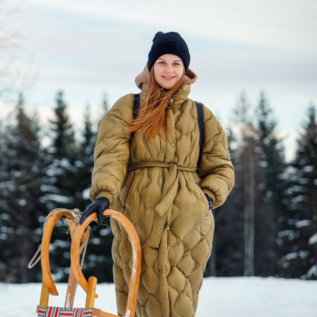A woman dressed warmly in a beige winter coat, black beanie, and gloves, holding a wooden sled outdoors in a snowy forest setting.

