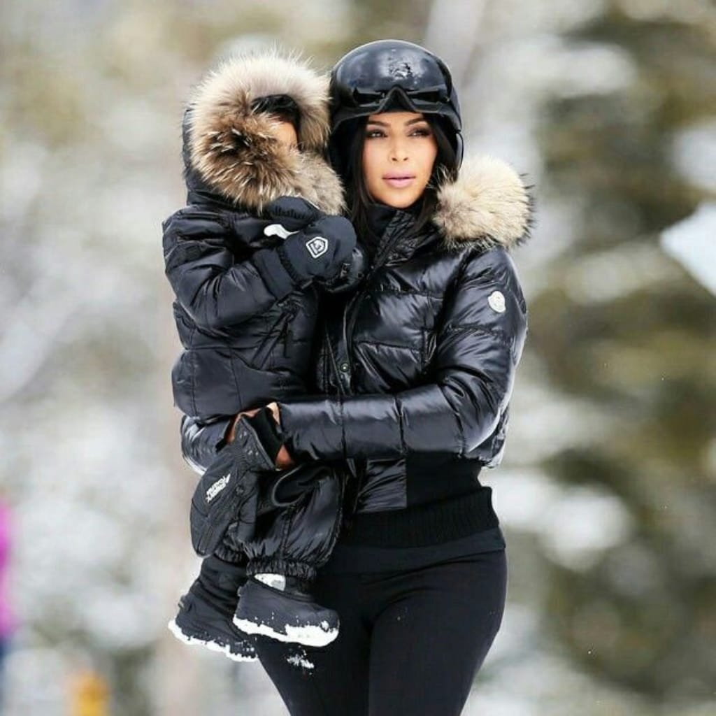 A woman dressed in a shiny black winter jacket with a fur-lined hood, holding a small child also bundled in a winter jacket with a fur-lined hood, outdoors in a snowy setting. The woman is wearing a black helmet and gloves, and the child is wearing snow boots.

