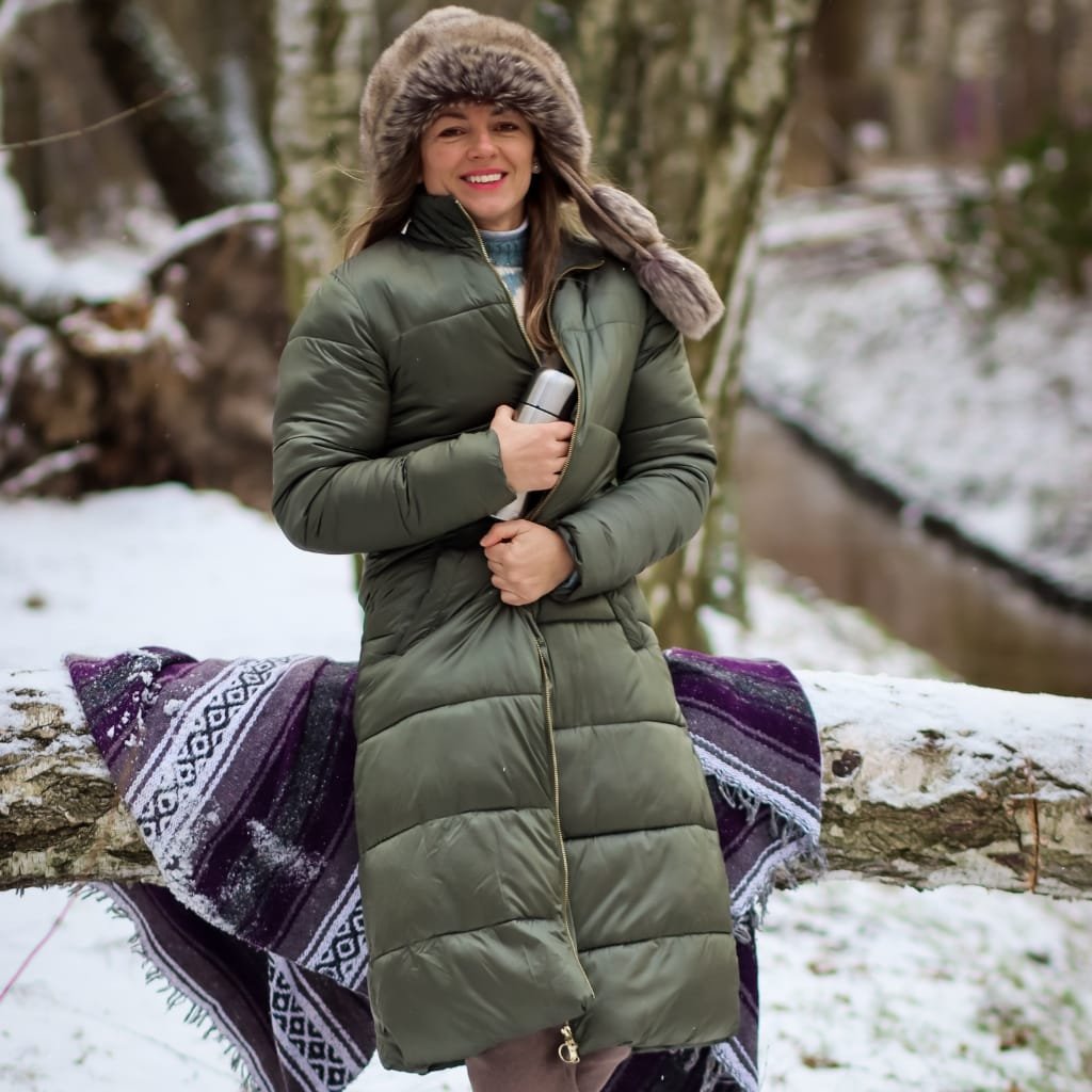  Woman standing outdoors in a snowy forest, wearing a green insulated winter coat with a fur-lined hood, holding a thermos, smiling, with a patterned blanket draped over a fallen log nearby.