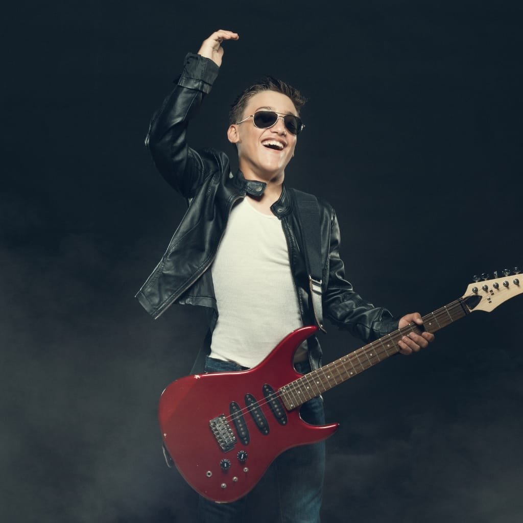 A young man wearing a black leather biker jacket, sunglasses, and a white T-shirt, holding a red electric guitar, smiling and gesturing enthusiastically.

