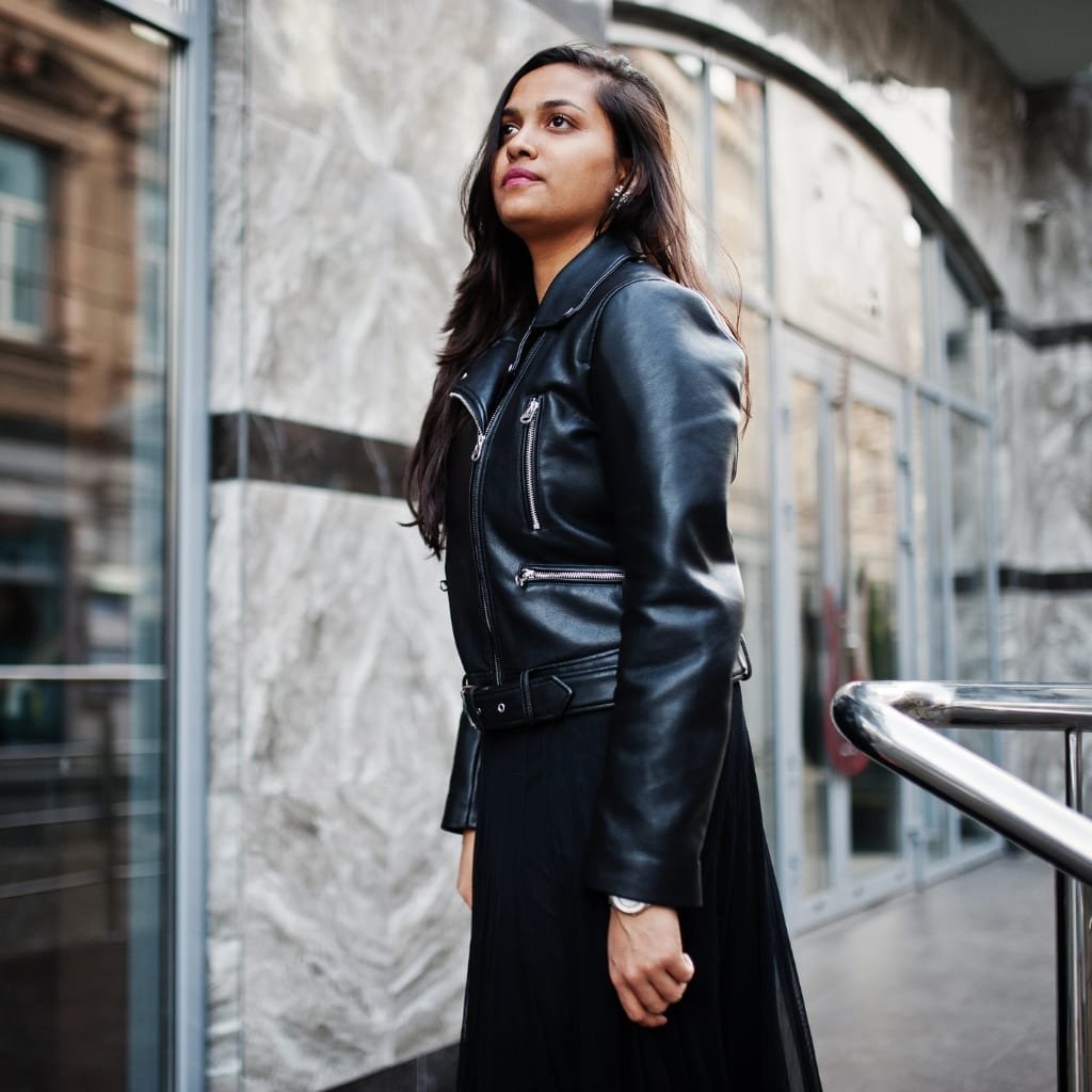 A stylish woman wearing a black leather jacket with silver zippers, standing outdoors against a modern, marble-clad building.