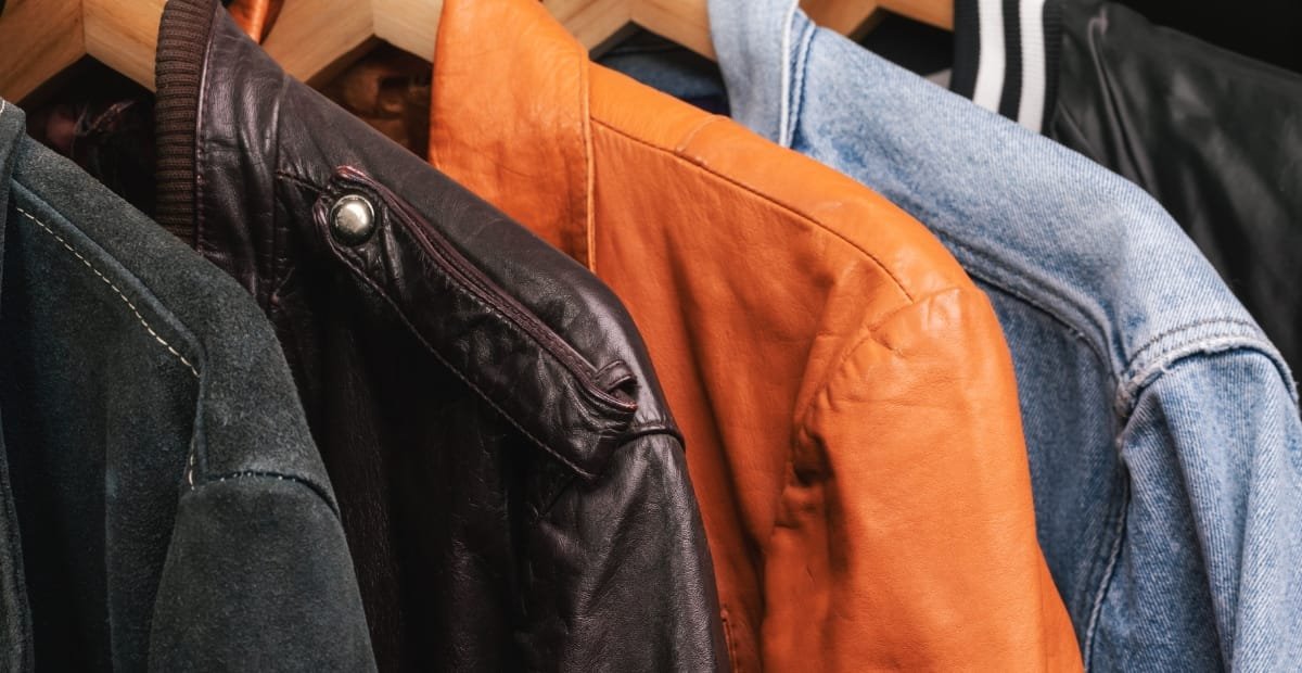Close-up image of four different leather jackets hanging on a rack, featuring various styles and colors such as black, brown, orange, and light blue denim.