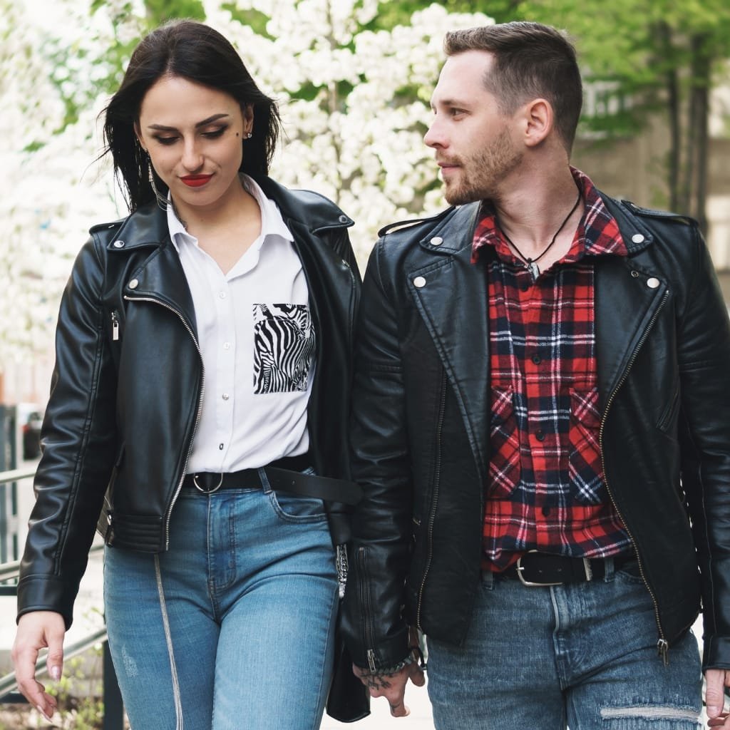 A man and a woman walking outdoors hand-in-hand, both wearing black leather shirt jackets. The woman has long dark hair, a white shirt with a zebra print pocket, and blue jeans. The man has short hair, a red plaid shirt, and distressed jeans.

