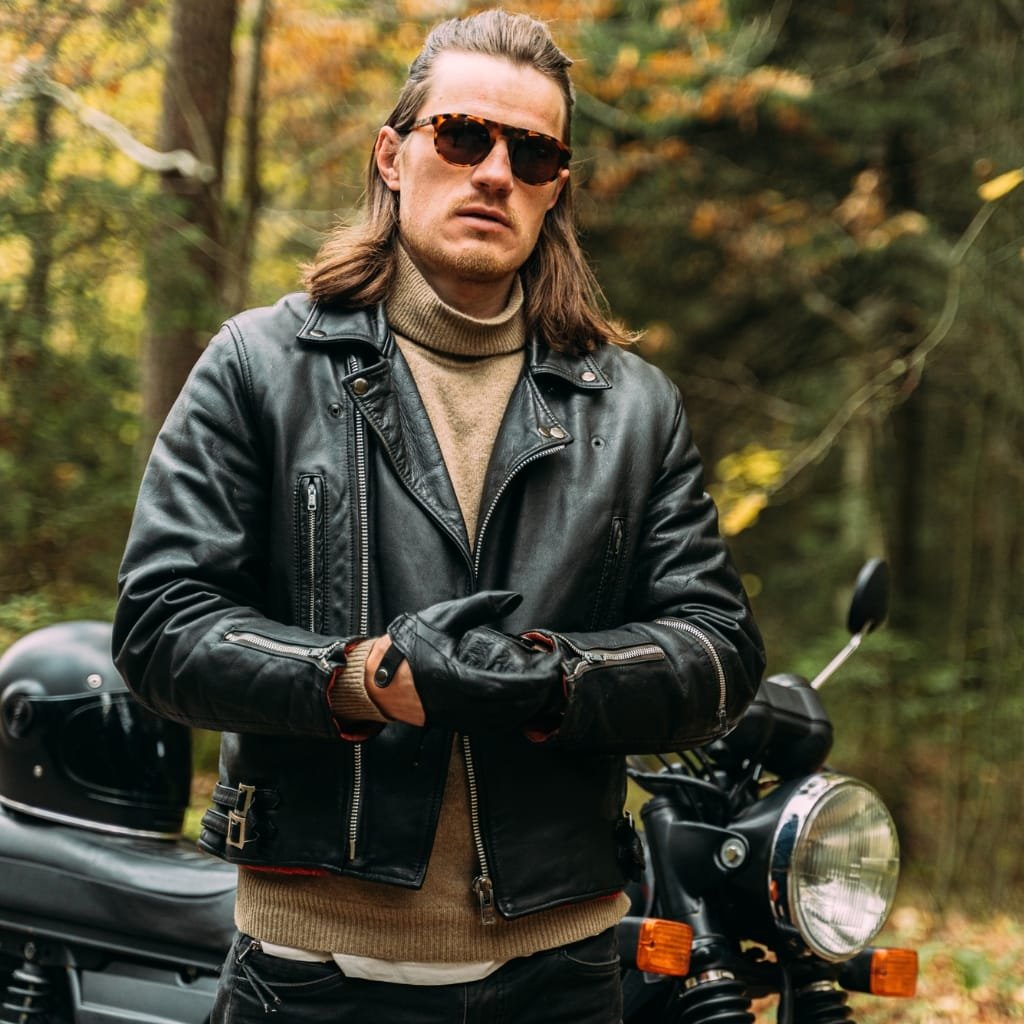 A man wearing a black leather biker jacket, sunglasses, and gloves standing in a forest with a motorcycle in the background.


