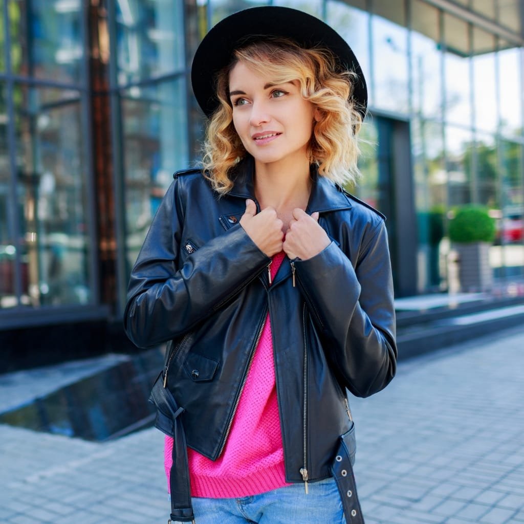 A young woman with blonde curly hair wearing a black wide-brim hat, a dark blue leather jacket, and a hot pink sweater, standing outdoors near a modern glass building.
