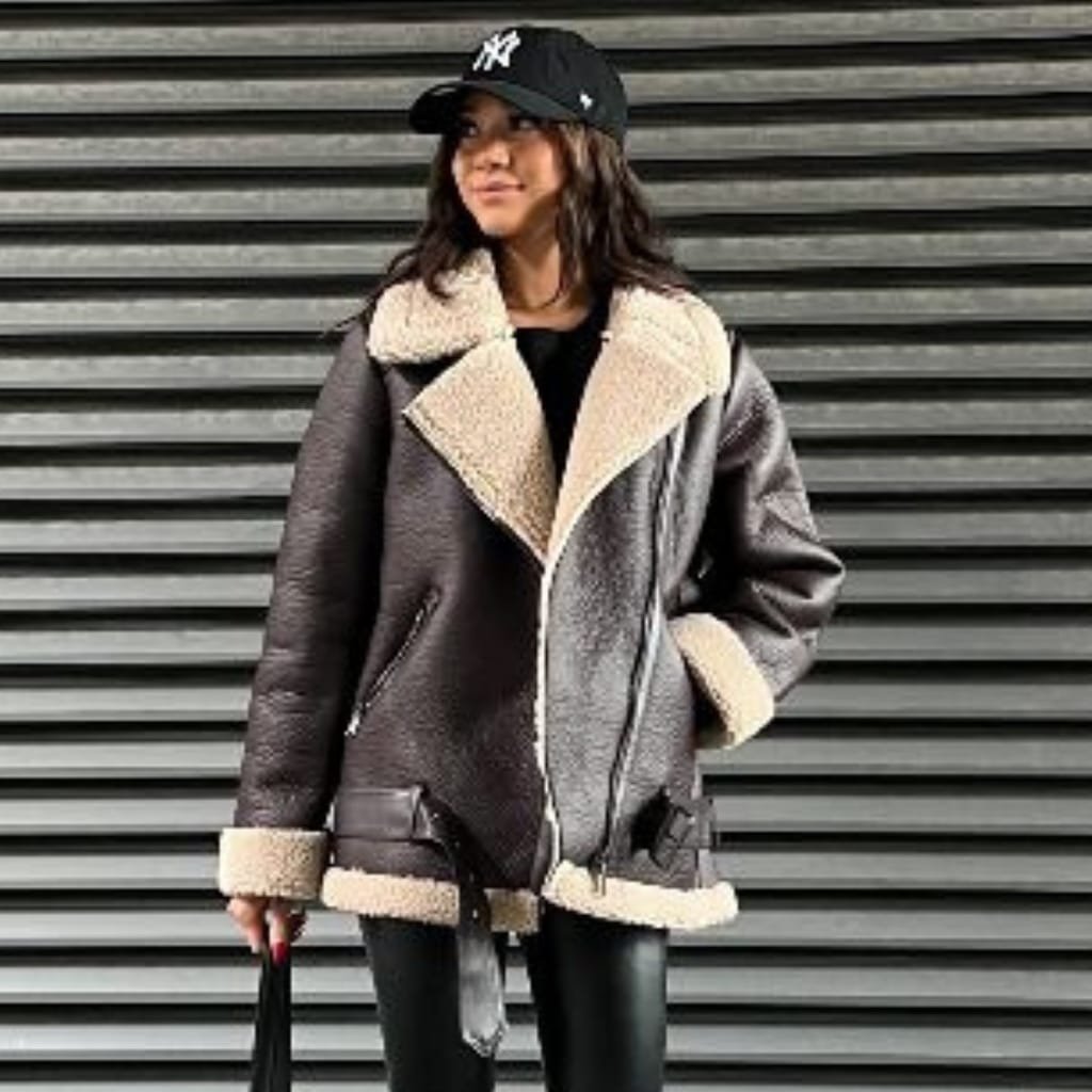 A woman standing in front of a metallic horizontal-slatted wall, wearing a dark brown shearling aviator jacket with cream-colored lining, a black baseball cap, black pants, and holding a small bag.

