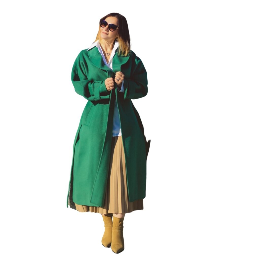 A woman wearing a long green windbreaker jacket over a white shirt, paired with beige pleated skirt, yellow boots, and sunglasses.