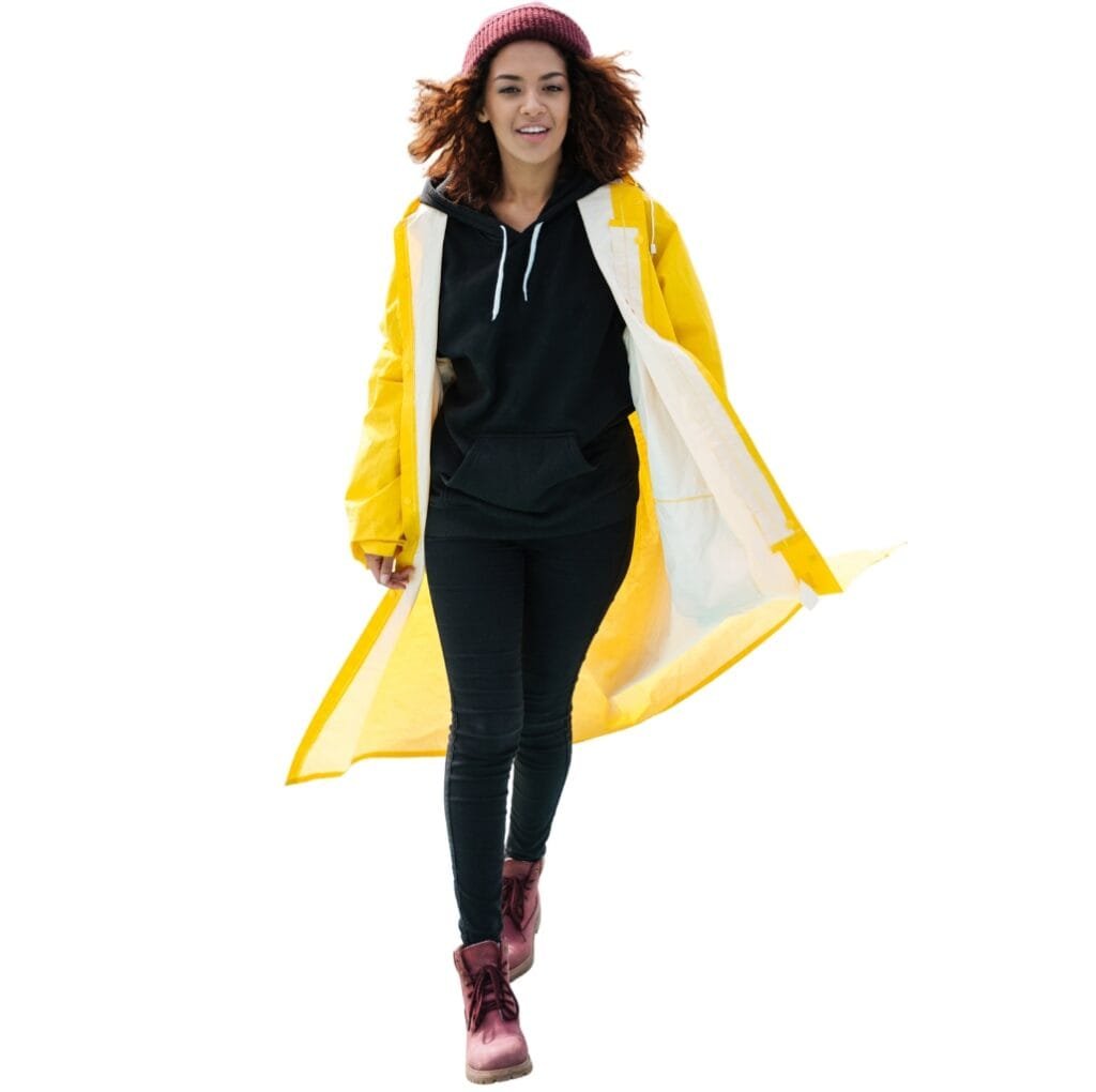 A young woman walking outdoors, wearing a vibrant yellow long windbreaker jacket over a black hoodie, paired with black skinny jeans and pink lace-up boots. She has curly hair, a beanie, and an energetic smile.