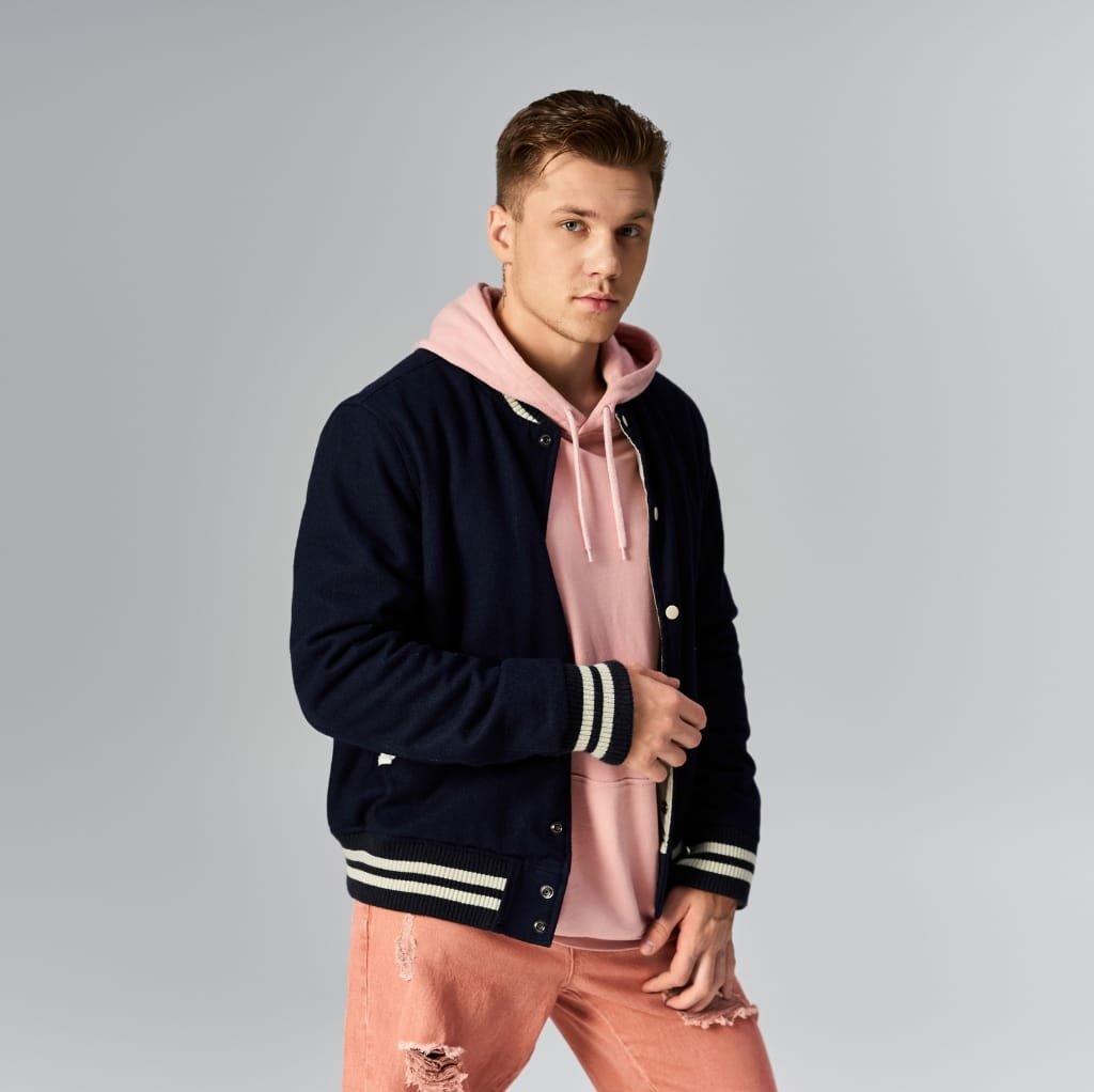 A young man with light brown hair, wearing a navy blue varsity jacket with white striped cuffs and collar, layered over a pink hoodie, and distressed pink jeans, standing against a plain grey background.