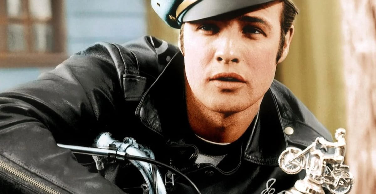 Marlon Brando: The Man Who Made the Leather Jacket Iconic A close-up image of a young man in a black leather biker jacket and a white helmet, sitting on a motorcycle. He has a serious expression and is looking slightly to the side. The background is slightly blurred.