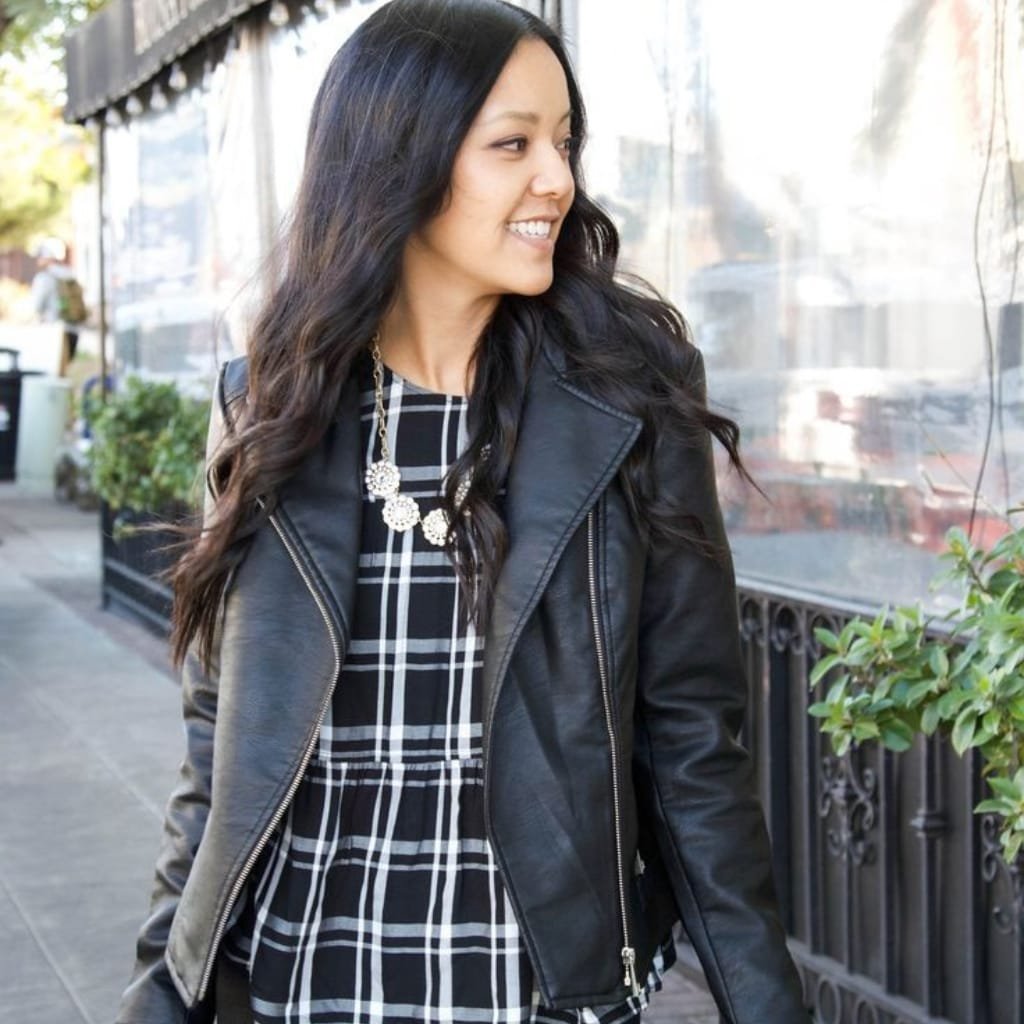 A woman wearing a black leather jacket over a black and white plaid dress, walking outdoors on a city sidewalk. She has long dark hair and is smiling, accessorized with a statement necklace.

