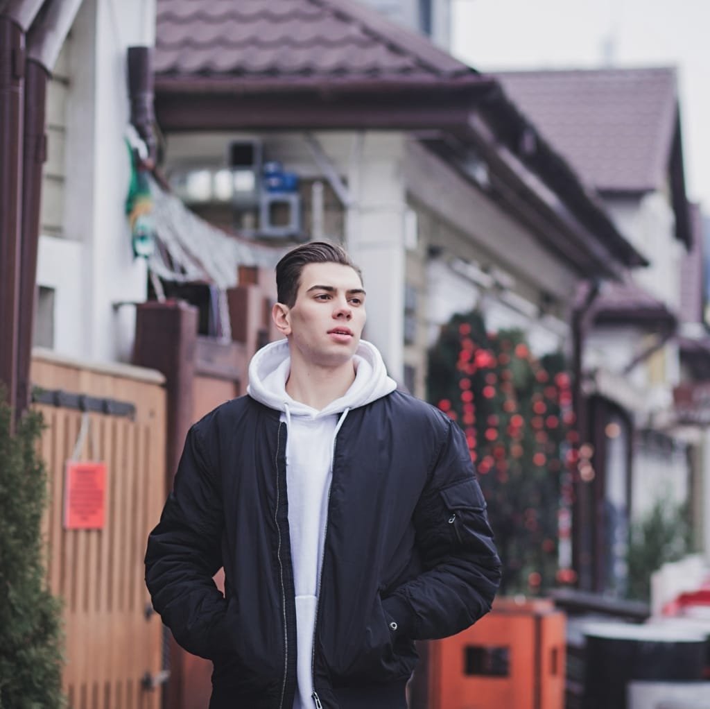 A young man walking outdoors wearing a minimalist black bomber jacket layered over a white hoodie, with a residential street background.