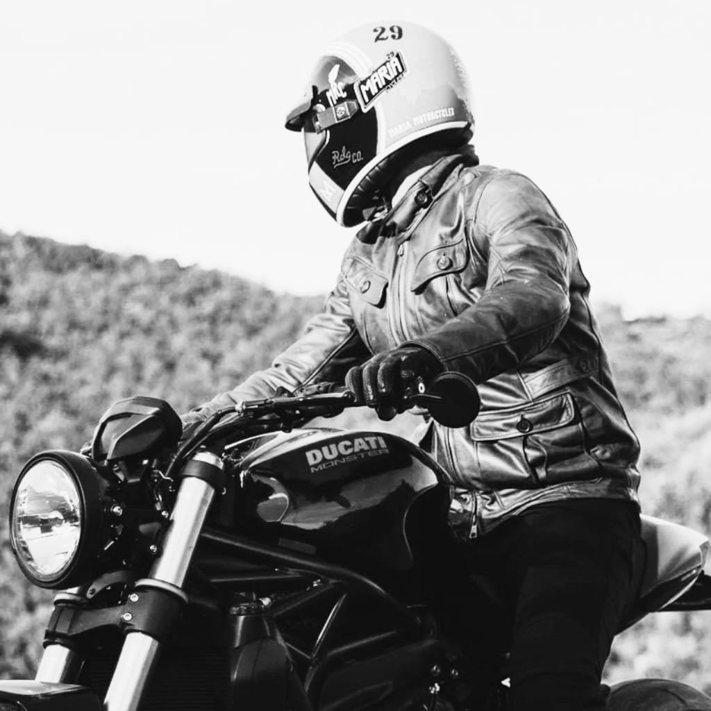 A black and white photo of a person riding a Ducati Monster motorcycle outdoors. The rider is wearing a leather jacket, gloves, and a helmet with various stickers, including what appears to be the name "Maria." The background is blurred, highlighting the rider and motorcycle.
